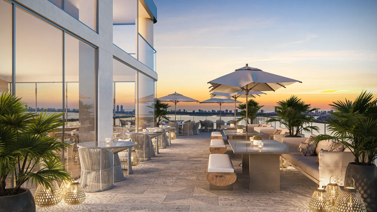 Rooftop terrace with shaded dining tables, lanterns and palms overlooking the water at sunset