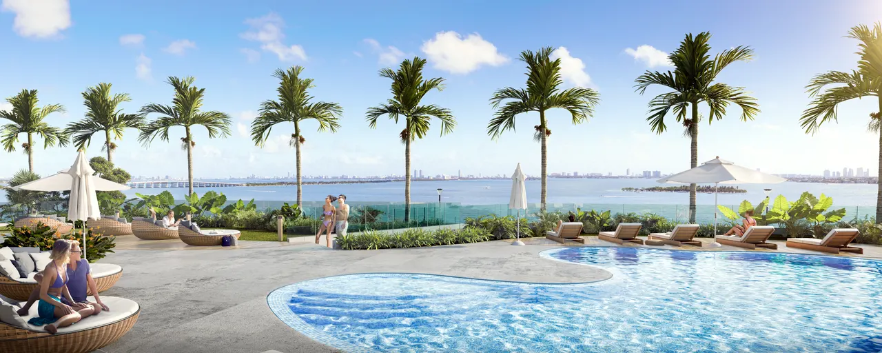 Curved resort pool with sun loungers, umbrellas and palm trees facing the water and skyline