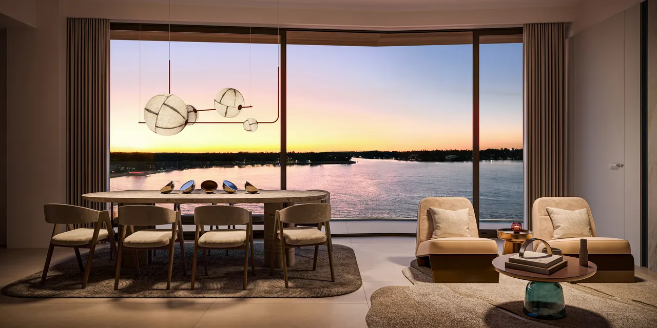 Dining area and lounge chairs beside floor-to-ceiling windows with a water view at sunset