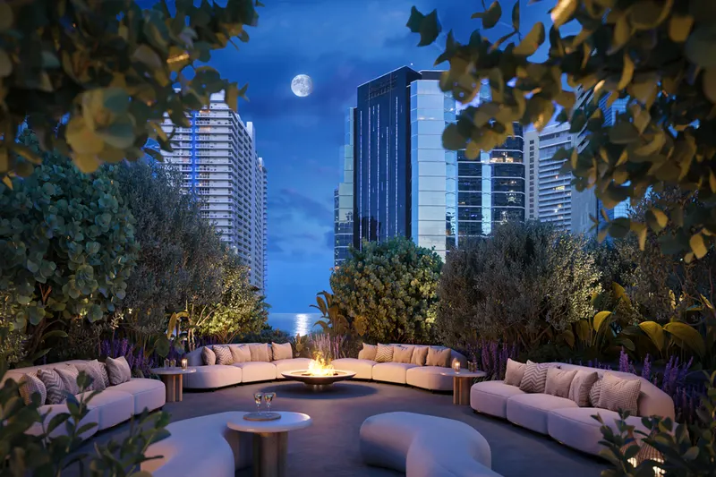 Landscaped rooftop terrace at dusk with curved sofas around a fire pit and city towers beyond