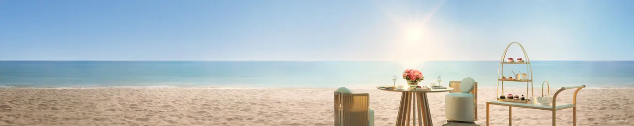 Beachfront table set for tea service with woven chairs and a tiered cart on open sand